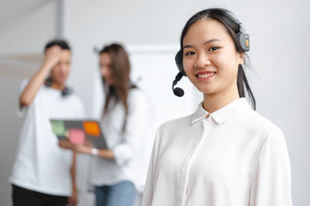 pexels photo 7681581 Young woman in a call center with colleagues in the background, smiling and wearing headphones.